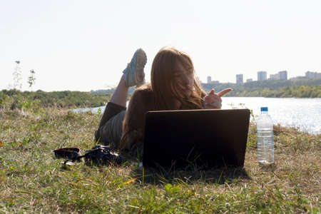 A girl with a laptop is lying on the river bank in a park outside the city. The concept of...