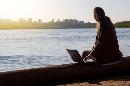 A girl with a laptop is sitting on a log by the river bank in a park outside the city. Concept of freelance, remote work or training. An independent female businessman works in nature in traveling.の写真素材