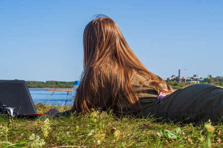 A girl with a laptop is lying on the river bank in a park outside the city. The concept of freelance, remote work or training. An independent female businessman works in nature while traveling.の写真素材