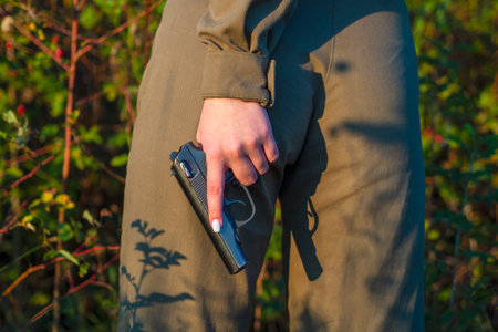 A girl in a green military jumpsuit stands against the background of red bushes and rosehip berries in the forest. A woman soldier holds, pulls out a police or army pistol in her hand at body level.の写真素材