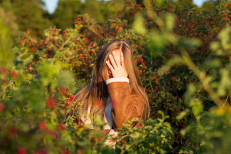 A bright, warm autumn portrait of a young Caucasian girl in a leather sheepskin coat against a background of red bushes and rosehip berries. The woman shows a gesture, a facepalm sign.の写真素材