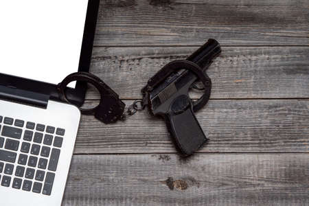 Laptop against the background of a gray wooden table next to a black police pistol in handcuffs. Flat lay, top view. The concept of Internet security and online crime.の写真素材