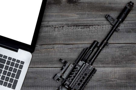 Laptop against the background of a gray wooden table next to a black army rifle. Flat lay, top view. The concept of Internet security and online crime.の写真素材