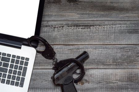 Laptop against the background of a gray wooden table next to a black police pistol in handcuffs. Flat lay, top view. The concept of Internet security and online crime.の写真素材