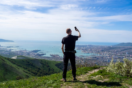 Man in a black T-shirt, glasses takes a selfie (photographed) in the mountains against the background of the city. Green hills and sea port. Novorossiysk, Krasnodar Territory, Russia.の写真素材