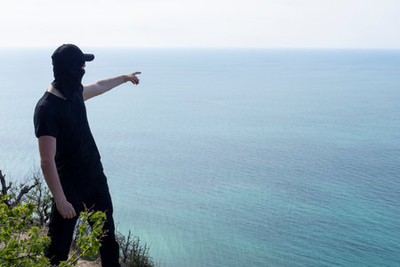 Man in a black cap, T-shirt and balaclava from the coastal mountains points his hand (finger) into the distance at the sea. Novorossiysk, Krasnodar Territory, Russia.の写真素材