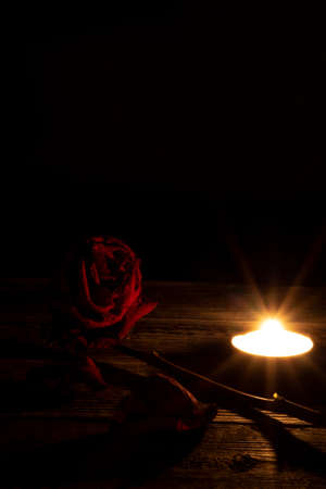 One red dried rose, dry petals next to a tea candle on a wooden table on a black, dark background. Dramatic mystical atmosphere black magic ritual. The concept of grief and mourning postcard.の写真素材
