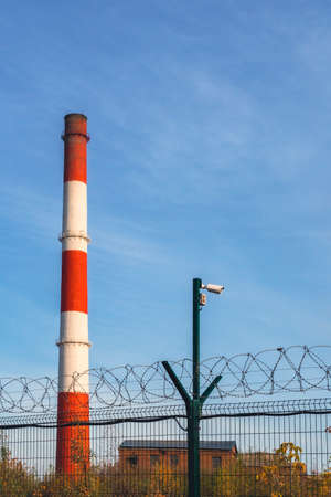 Fence with barbed wire on the background of smoking red and white pipes at a thermal power plant...