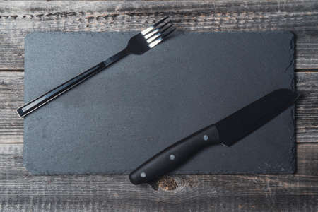 An empty black cutting slate board with a serving fork and knife on a wooden table background. Copy space is the place to insert.の写真素材