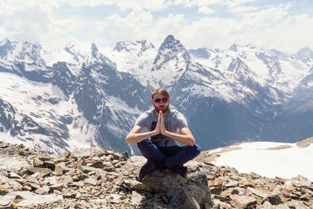 A hipster man with sunglasses and a beard in a gray T-shirt sits and meditates against the background of snowy rocks. Caucasus Mountains, Dombay. The concept of harmony and inner balance.の写真素材