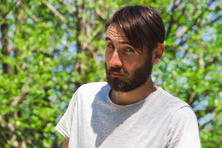 Portrait of a surprised young Caucasian man in close-up. Bearded hipster guy with bangs on the background of green foliage of trees in the park.の写真素材