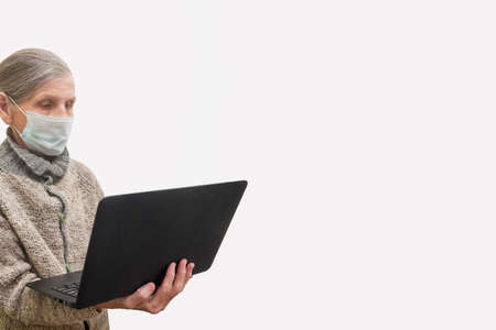 An elderly woman in a medical mask and a knitted sweater is holding a black laptop on a white...