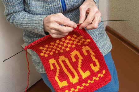 Lonely old elderly woman is knitting needle a winter New Year's Christmas scarf with the numbers 2023 from red and white yarn. The concept of a retired hobby, creative handmade.の写真素材