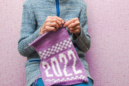 Lonely old elderly woman is knitting needle a winter New Year's Christmas scarf with the numbers 2022 from pink and white yarn. The concept of a retired hobby, creative handmade.の写真素材