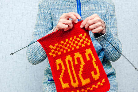 Lonely old elderly woman is knitting needle a winter New Year's Christmas scarf with the numbers 2023 from red and white yarn. The concept of a retired hobby, creative handmade.の写真素材