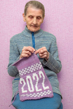 Lonely old elderly woman is knitting needle a winter New Year's Christmas scarf with the numbers 2022 from pink and white yarn. The concept of a retired hobby, creative handmade.の写真素材