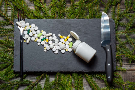Pills scattered on a black slate board lie next to a serving fork and knife on a Christmas background of fir branches. Diet and dietary supplements. Concept of poisoning treatment after the new year.の写真素材