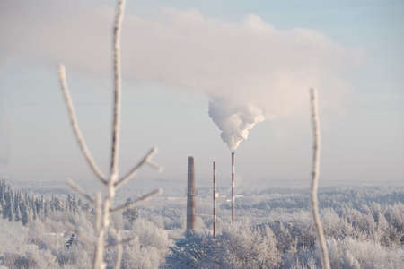 Winter landscape. A forest covered with snow, a factory with a pipe releasing smoke on the background. Heating season. The concept of environmental pollution.の写真素材