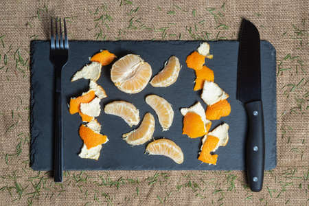 Pieces of peeled tangerine and peel lie on a black slate board next to a serving knife and fork...