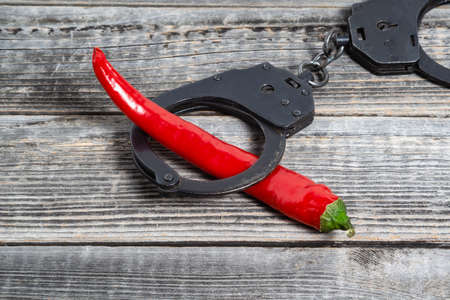 A red hot chili pepper lies shackled in black police handcuffs against the background of a gray...