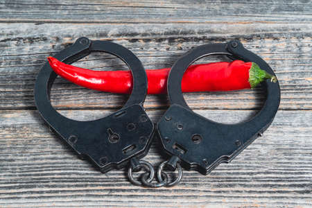 A red hot chili pepper lies shackled in black police handcuffs against the background of a gray wooden table. The concept of sexual crimes. Punishment for violence and harassment.の写真素材