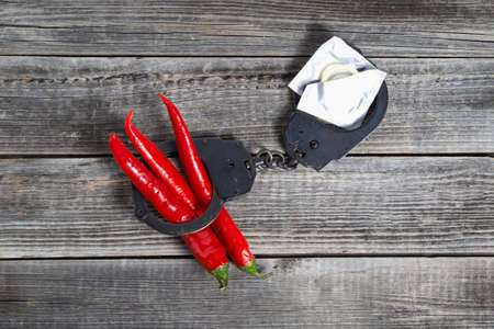 Three red hot chili peppers lie shackled in black police handcuffs with condoms on a gray wooden table background. The concept of sexual crimes. Punishment for prostitution.の写真素材