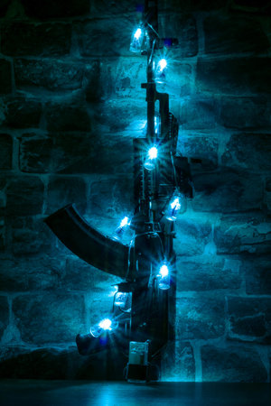 Assault automatic rifle in the form of a Christmas tree decorated with an electric garland of vials on the background of a gray brick wall in the night room.の写真素材