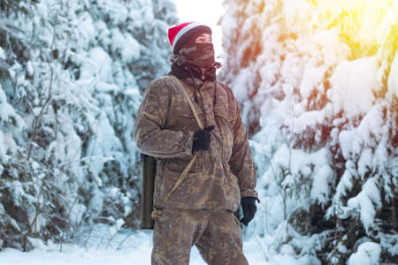 A military man in camouflage is holding a rocket launcher in the outdoors. A soldier with a gun in the winter forest. A male in a red Christmas hat.の写真素材