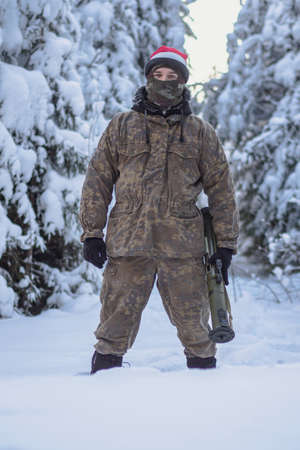 A military man in camouflage is holding a rocket launcher in the outdoors. A soldier with a gun in the winter forest. A male in a red Christmas hat.の写真素材