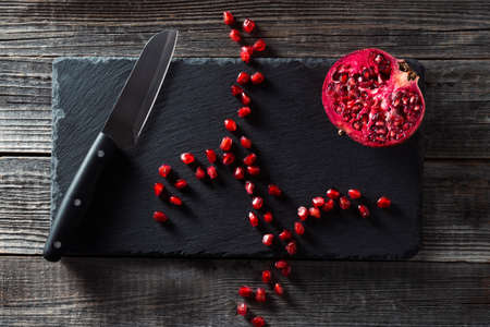 Half a red pomegranate fruit and a pile of seeds on a black slate cutting board with a kitchen knife on a wooden table background. The concept of healthy eating, diet.の写真素材