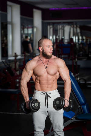 Man with a naked torso poses the background of a fitness room. Bodybuilder holds in his hands and trains with dumbbells in the gym. Pumped up guy show muscles. Concept of an active healthy lifestyleの写真素材