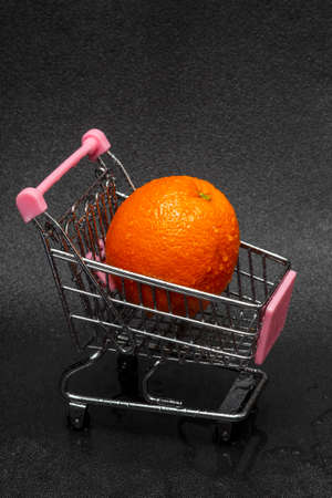 A washed orange with water drops in a shopping cart on a black background. The concept of selling or buying fruit.の写真素材