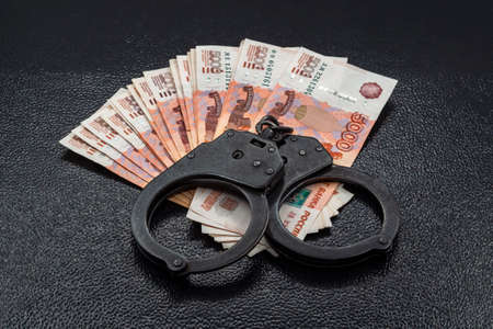 Many five thousand Russian money banknotes lie in a fan on a wooden table background with black police handcuffs. The concept of economic crime, theft or robbery.の写真素材