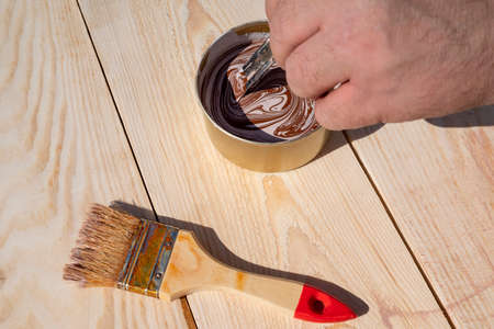 Man with his hand mixes brown paint in a tin can on wooden boards next to the brush. The concept of renovation, construction and repair.の写真素材