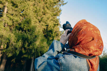 Elderly woman holds a camera in her hands against the background of a sunlight forest. Old lady takes aim at the viewfinder and takes a photo. Grandma photographs nature on a travel.の写真素材