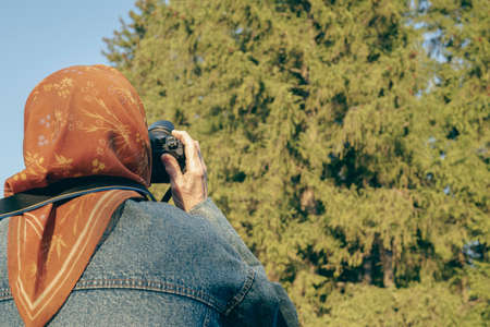Elderly woman holds a camera in her hands against the background of a sunlight forest. Old lady takes aim at the viewfinder and takes a photo. Grandma photographs nature on a travel.の写真素材