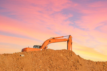 An orange excavator stands on an earthen slide against the background of the sunset sky. The concept...