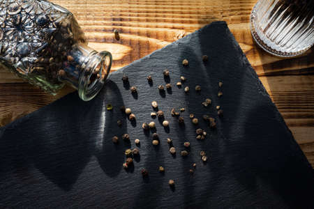 Mixture of peppers spilled from a glass bottle for storing spices on a black slate cutting board on...