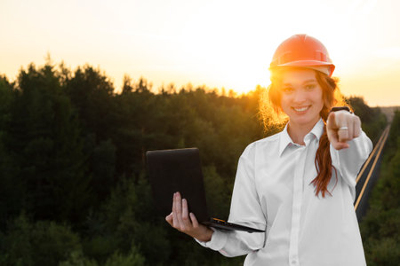 A female construction inspector in an orange helmet with red hair holds a laptop in her hands...
