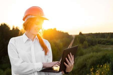 A female construction inspector in an orange helmet with red hair holds a laptop in her hands...