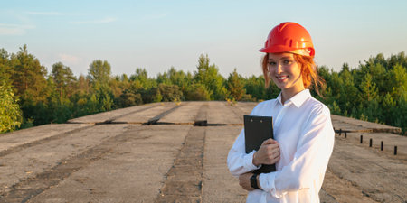 A female construction inspector in an orange helmet with red hair holds a tablet in her hands against the background of the bridge. Restoration of an abandoned object.の写真素材