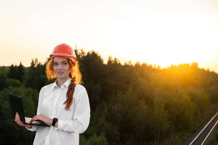 A female construction inspector in an orange helmet with red hair holds a laptop in her hands against the background of the bridge. Restoration of an abandoned object.の写真素材