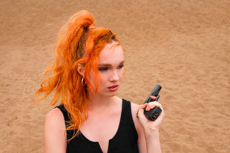 Portrait of a serious insidious secret agent girl in a black dress with red hair with a gun in her...