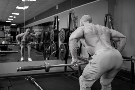 Man admires his reflection in the mirror of the fitness room. Bodybuilder holds in his hands and trains with barbell in the gym. Pumped up guy show muscles. black and white photography.の写真素材