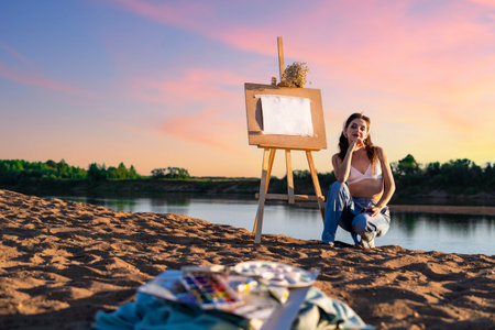 A woman during the open air on the sandy bank of the river with an easel. An artist with paints near an empty mock up sheet in nature.の写真素材
