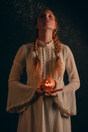 Woman stands and holds a small Jack's pumpkin lamp in her hands on a black dark background in a...