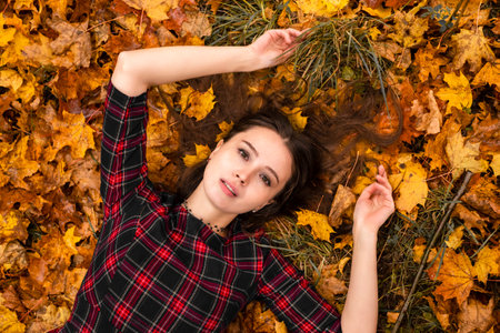 A sensual woman in a red plaid dress lies in autumn maple yellow leaves. The girl is resting in the...