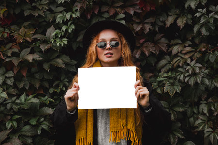 An outdoor girl in autumn clothes holds an empty sheet of paper in her hands while standing against a background of foliage. A woman with a mock up.の写真素材