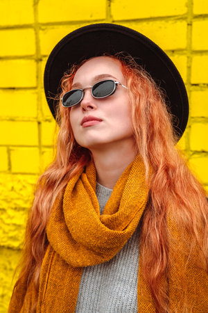 Portrait of a girl in a black round felt hat and sunglasses with an orange scarf. Female autumn city portrait on the background of a yellow brick wall.の写真素材