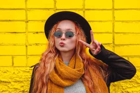 Portrait of a girl kissing lips in a black round felt hat and sunglasses with an orange scarf. Female autumn city portrait on the background of a yellow brick wall.の写真素材